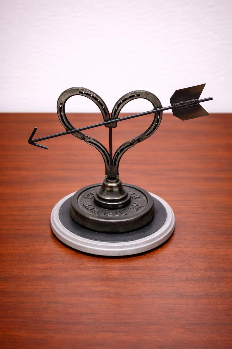 May include: A metal sculpture featuring a heart shape formed from two horseshoes, pierced by an arrow. The sculpture is mounted on a circular base with a dark gray top and a light gray rim. The arrow and heart are dark gray.