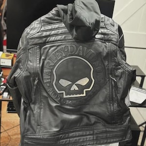 May include: Black leather motorcycle jacket with a hood. The back features a silver Harley-Davidson skull logo and the words "Harley-Davidson Motorcycles" in a circular design. The jacket has a quilted design on the shoulders and upper arms.