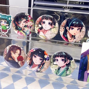 Puede incluir: Una colección de chapas redondas con ilustraciones coloridas de estilo anime de personajes con cabello oscuro y ojos expresivos, exhibidas en un soporte de acrílico transparente de varios niveles. Los diseños incluyen detalles florales y texto japonés.
