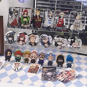 May include: A collection of anime-themed merchandise including rectangular cards, circular pin-back buttons, and acrylic charms featuring various characters. The items display colourful illustrations of figures in diverse outfits, arranged on a clear tiered display stand.