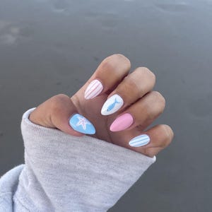 May include: Almond-shaped artificial nails with a coastal theme. The nails feature designs including pink and white stripes, a blue starfish, a white fish, and blue and white stripes. The nails are painted in pastel colours.