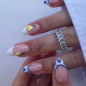 May include: Close-up of manicured nails with a French tip design. The nails feature a white tip with a yellow lemon design and a blue and white pattern. A silver ring with rectangular stones is also visible.