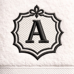 May include: White towel with an embroidered black monogram letter "A" inside a decorative frame. The frame has a scalloped edge and a stylized bow at the top. The background of the frame has a striped pattern.