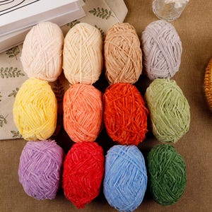 Puede incluir: Una colección de ovillos de lana coloridos en tonos rosa, amarillo, naranja, rojo, verde, morado, azul y marrón. La lana está dispuesta sobre una superficie marrón, con una cesta de mimbre y un libro en el fondo.