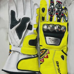 May include: Pair of motorcycle gloves in white and yellow leather. The gloves have black accents and protective metal plates. The right glove features the text "VR 46" and colorful decorative elements. The gloves are designed for motorcycle riding.