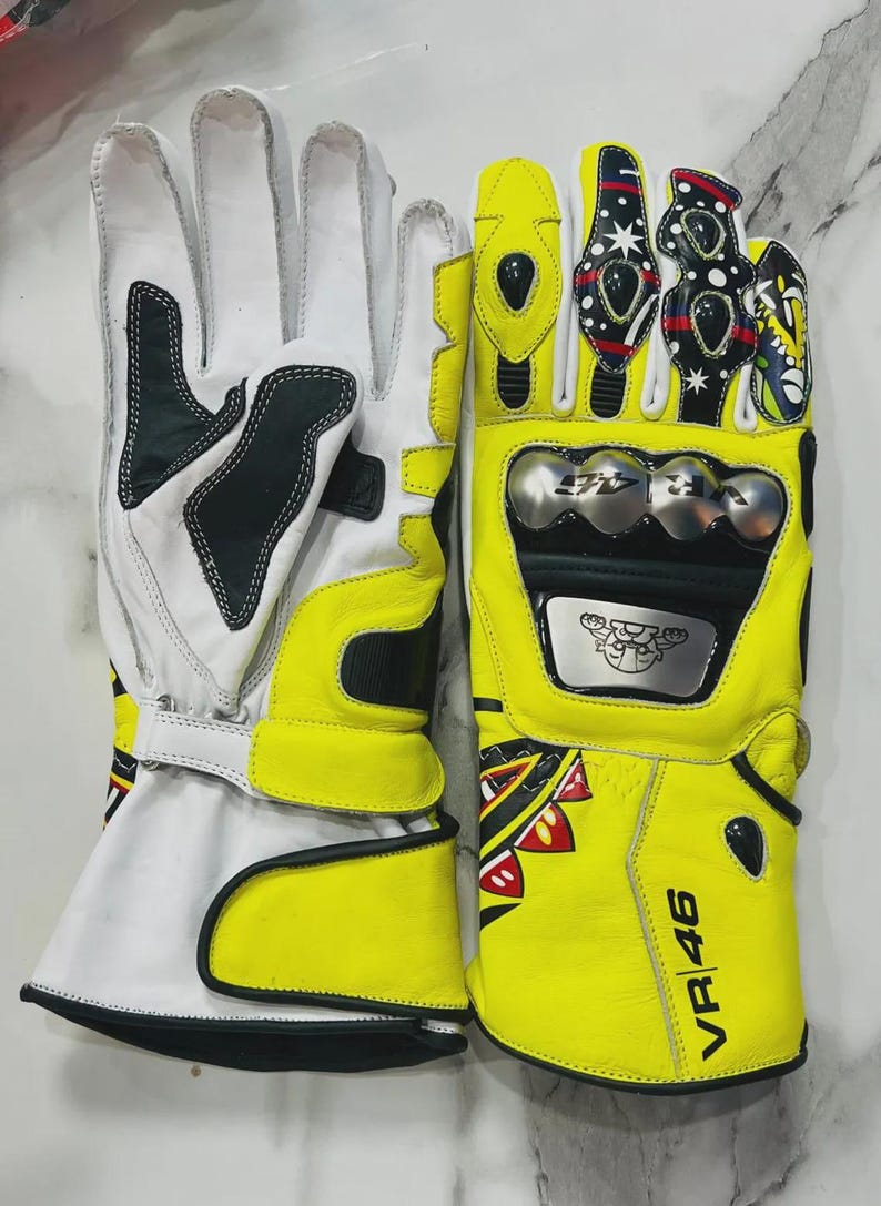 May include: Pair of motorcycle gloves in white and yellow leather. The gloves have black accents and protective metal plates. The right glove features the text "VR 46" and colorful decorative elements. The gloves are designed for motorcycle riding.