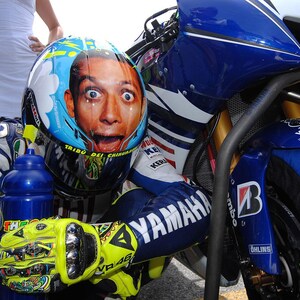 May include: A motorcycle racer wearing a blue, white, and yellow Yamaha racing suit and helmet. The helmet features a portrait of a surprised face. The motorcycle is blue and white with the number 46.