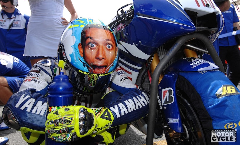 May include: A motorcycle racer wearing a blue, white, and yellow Yamaha racing suit and helmet. The helmet features a portrait of a surprised face. The motorcycle is blue and white with the number 46.