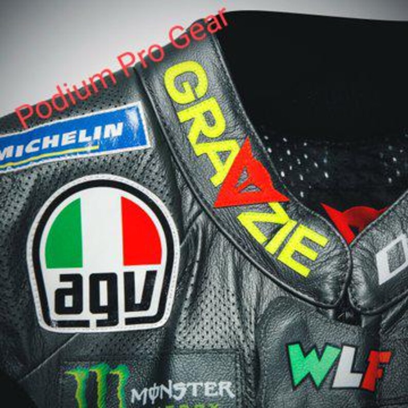 May include: A black leather motorcycle jacket featuring a perforated texture, a collar with the yellow text GRAZIE and a red logo, an AGV logo patch, a blue Michelin patch, a green Monster Energy logo, and the text WLF in green, white, and red.