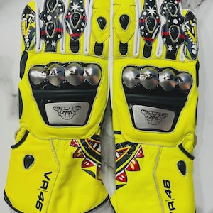May include: Pair of bright yellow motorcycle gloves with black and white accents. The gloves feature silver knuckle protectors and the text "VR 46". The design includes decorative patterns and logos.