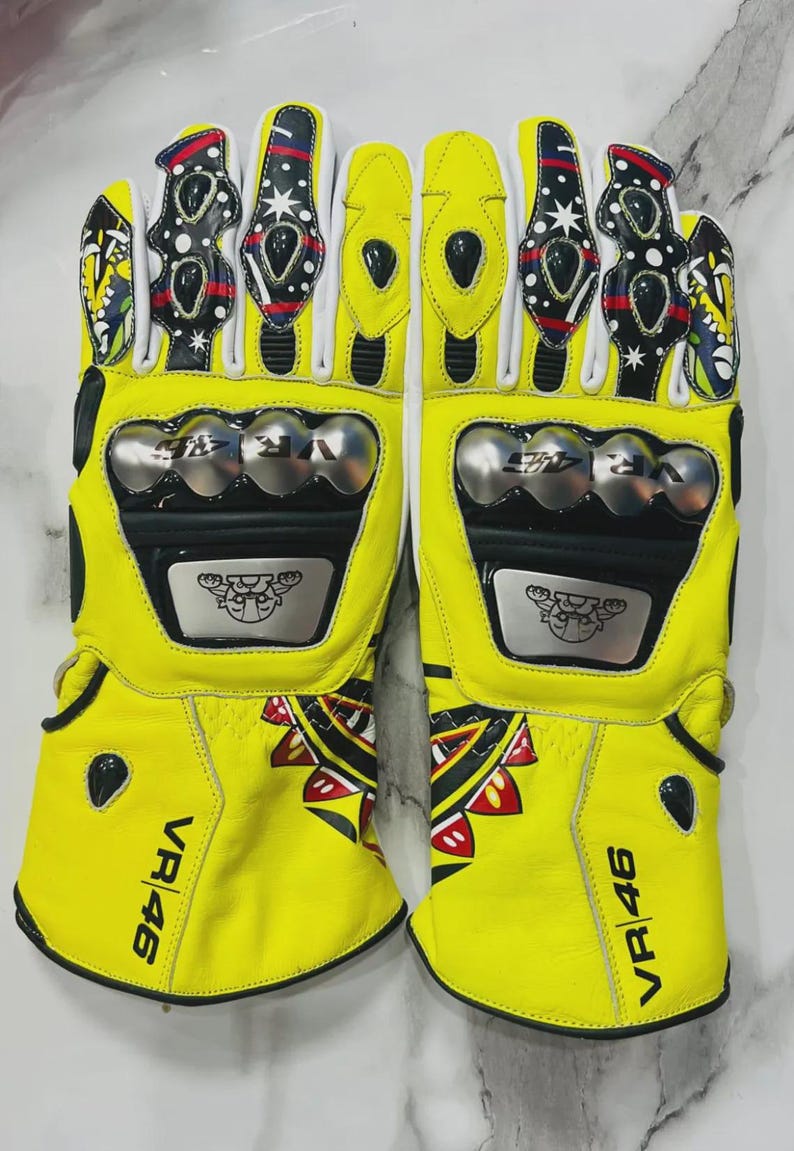 May include: Pair of bright yellow motorcycle gloves with black and white accents. The gloves feature silver knuckle protectors and the text "VR 46". The design includes decorative patterns and logos.