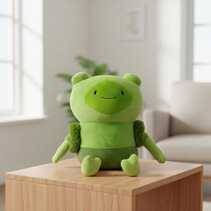 May include: A plush toy in shades of green, resembling a cartoon character. The toy has a round head with small ears, a smiling face, and a body with arms and feet. It is sitting on a wooden surface, with a blurred background of a window and a couch.