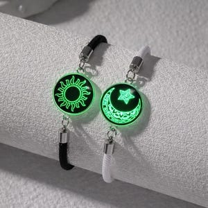 Peut inclure: Deux bracelets assortis avec des pendentifs soleil et lune verts brillants. Un bracelet a un cordon noir, l'autre un cordon blanc. Les pendentifs sont ronds avec des accents métalliques argentés.