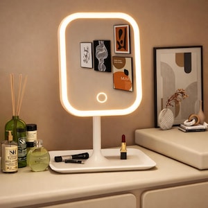 Puede incluir: Un espejo de tocador blanco e iluminado con un marco rectangular y una luz circular. El espejo tiene una bandeja incorporada que contiene artículos de maquillaje. Pequeñas impresiones artísticas están adheridas a la superficie del espejo.