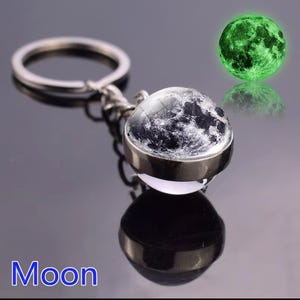 Peut inclure: Un porte-clés en métal argenté doté d'un pendentif sphérique en verre avec un graphique détaillé de la surface lunaire en noir et blanc. Le pendentif est conçu pour briller dans l'obscurité, émettant une lumière verte, fixé à un anneau.