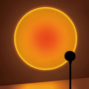 May include: A sunset projection lamp featuring a black circular head on a thin vertical stand, casting a large, vibrant, glowing orange and yellow circular light pattern that resembles a sun against a flat, neutral-toned wall surface.