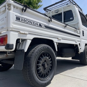 Puede incluir: Mini camión Honda Acty blanco con ruedas y neumáticos negros. El camión tiene una baca negra y el logotipo de Honda es visible en el lateral. El camión está estacionado sobre una superficie pavimentada.