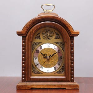 Peut inclure: Une horloge à quartz en bois avec un cadran doré et des chiffres romains. L'horloge a une poignée sur le dessus et les mots "Tempus Fugit" sont visibles sur le cadran. L'horloge est rectangulaire avec un dessus en arc et repose sur une base en bois.