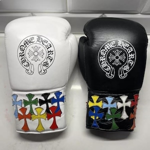 Puede incluir: Un par de guantes de boxeo, uno blanco y otro negro. Cada guante presenta un emblema circular con texto y un diseño de cruz. Las muñequeras están adornadas con patrones de cruces coloridas. Los guantes se muestran sobre un fondo de baldosas blancas.
