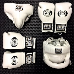 Puede incluir: Conjunto de equipo de boxeo blanco, que incluye guantes, casco y protector inguinal. Cada pieza presenta el logotipo "REYES" en negro y dorado. Los artículos están dispuestos sobre una superficie negra texturizada, mostrando equipo de boxeo profesional.