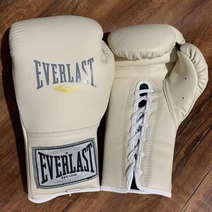 May include: Cream-coloured boxing gloves with the "EVERLAST" logo in grey and yellow. The gloves are made of leather and have white laces. The gloves are on a wooden surface.
