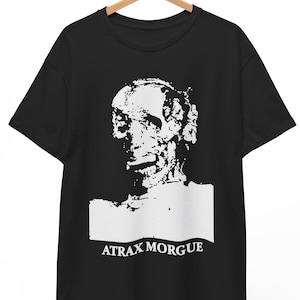 May include: A black short-sleeved t-shirt featuring a high-contrast white graphic print of a human skull and facial features. Below the graphic, the text ATRAX MORGUE is printed in a bold, white serif font. The shirt is displayed on a wooden hanger.