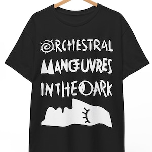Może przedstawiać: Czarny t-shirt z krótkim rękawem z białym napisem graficznym ORCHESTRAL MANOEUVRES IN THE DARK. Pod tekstem znajduje się biała, stylizowana sylwetka twarzy z profilu z zamkniętym okiem i długimi rzęsami, nadrukowana na przodzie koszulki.