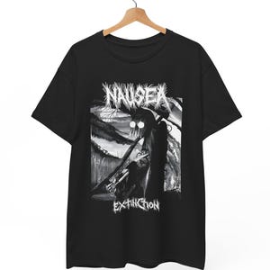 May include: Black t-shirt with a monochrome graphic. The design features the word "NAUSEA" above an illustration of a figure with glowing eyes, and the word "EXTINCTION" below. The t-shirt is suspended from a wooden hanger.