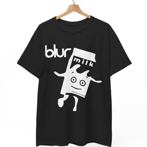 May include: Black t-shirt with the word "blur" in white, above a cartoon milk carton character. The milk carton has a smiling face and the word "milk" on it. The word "pasteurised" is below the milk carton.
