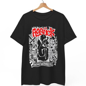 May include: A black short-sleeve t-shirt featuring a white graphic print with red text that reads "ABBIICK" at the top and "BORN BACKWARDS" at the bottom. The design includes a central figure, a dollar sign, and various abstract sketches.