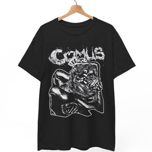 May include: Black t-shirt with a white graphic. The graphic features the word "COMUS" in a distressed font above a detailed illustration of a figure with a distorted face and body. The shirt is hanging on a wooden hanger.