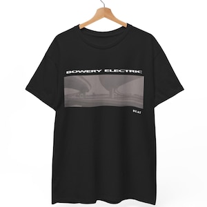 以下が含まれることがあります： 「BOWERY ELECTRIC」の文字と建物のグレースケール画像がプリントされた黒いTシャツ。画像の下には「BEAT」の文字。木製のハンガーに吊るされたTシャツ。