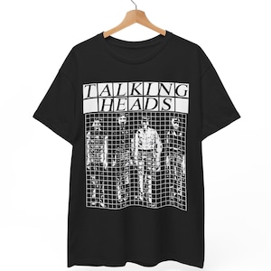 以下が含まれることがあります： 白いグラフィックプリントが施された黒の半袖Tシャツ。ブロックフォントで「TALKING HEADS」という文字が書かれており、その下に4人の人物が並んでいる白黒のイラストがグリッドパターンの中に描かれています。