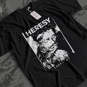 以下が含まれることがあります： 黒の半袖Tシャツ。顔の白いグラフィックプリントが施され、上部に「HERESY」、下部に「1985-'87」の文字がある。襟元には「L LARGE」と書かれた白い長方形のタグが細い白い紐で取り付けられている。