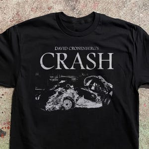Könnte beinhalten: Schwarzes T-Shirt mit dem weißen Text "DAVID CRONENBERG'S CRASH" über einem Graustufenbild von zwei Autowracks. Das T-Shirt hat einen Rundhalsausschnitt.