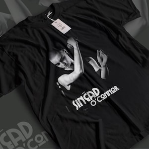 Op de afbeelding: Een zwart T-shirt met korte mouwen en een monochrome fotografische print van Sinéad O'Connor die een microfoon vasthoudt. De tekst "SINÉAD O'CONNOR" is in het wit onder de afbeelding gedrukt. Een wit maatlabel "L" is aan de kraag bevestigd.