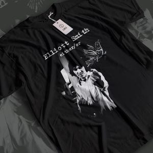May include: Black t-shirt featuring a grayscale image of a person pointing, with the text "Elliott Smith either/or" and a bird illustration. A white tag with the word "LARGE" is attached.