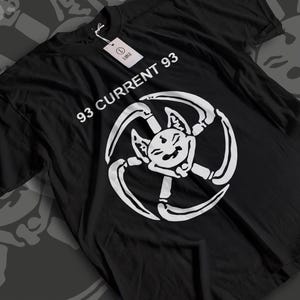 以下が含まれることがあります： 黒いTシャツ。白で「93 CURRENT 93」と書かれています。シャツには、曲がった付属肢を持つ、笑顔の猫のような生き物の白いグラフィックが描かれています。白いタグがシャツに付いています。