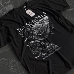 May include: Black t-shirt featuring a white graphic of ammonite fossils. The text "The Sundays" is printed above the fossils, with "Reading, Writing and Arithmetic" below. A white tag indicates the size is large.