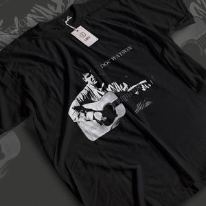 May include: A black short-sleeved t-shirt featuring a white graphic print of Doc Watson playing an acoustic guitar. The text "DOC WATSON" is printed above the graphic. A white rectangular tag labelled "L LARGE" is attached to the collar.