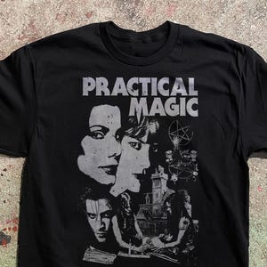 Puede incluir: Una camiseta negra de manga corta con un estampado gráfico gris desgastado. El diseño incluye el texto "PRACTICAL MAGIC" sobre los retratos de dos personas, un símbolo de pentagrama y una ilustración detallada de una casa grande.