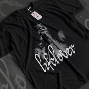 May include: Black t-shirt featuring a grayscale graphic of a person holding a microphone, with the word "lifelover" in white script. A white tag with the word "LARGE" is attached to the shirt.