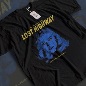 以下が含まれることがあります： 黒いTシャツ。黄色で「LOST HIGHWAY」の文字、女性の顔の青と黄色のグラフィック。タイトル上には「DIRECTED BY DAVID LYNCH」の文字。グラフィックの下には「...DO YOU OWN A VIDEO CAMERA?」のフレーズ。