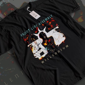 May include: Black t-shirt featuring the text "FRONT LINE ASSEMBLY" in teal and a graphic design with white and red accents. The word "Millennium" is printed in gold. A white tag with the word "LARGE" is attached.