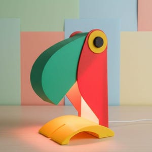 May include: A colorful toucan-shaped desk lamp. The lamp features a green beak, red body, and yellow base. The eye is a yellow circle with a black center. The lamp is plugged in and emits a warm glow.