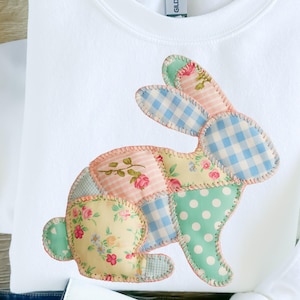 May include: White sweatshirt featuring a patchwork bunny design. The bunny is composed of various fabric patterns, including floral, polka dot, and gingham, in pastel colors. The design is stitched onto the front of the sweatshirt.