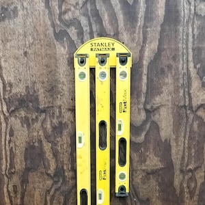 May include: A set of three yellow Stanley FatMax spirit levels of varying lengths, mounted on a shared arched bracket at the top. Each level features multiple integrated liquid vials for horizontal and vertical leveling, with black hand grips.