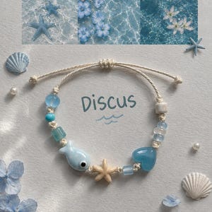 May include: A cream-coloured cord bracelet featuring a central beige starfish charm, a light blue fish bead with an eye, a blue heart bead, and various translucent blue and white beads. The word Discus is printed in blue above the bracelet.