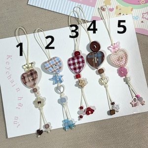 May include: Five handmade keychains with heart-shaped charms. Each keychain features a different patterned heart, adorned with beads, flowers, and other decorative elements. The keychains are attached to a white card with text.