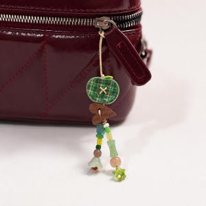 May include: A deep burgundy quilted bag with a zipper pull featuring a dangling charm. The charm consists of a green plaid apple-shaped button, a small wooden heart, and two strands of colorful glass beads in shades of yellow, green, and blue.